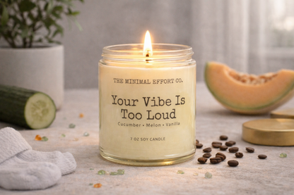 Candle labeled 'Your Vibe Is Too Loud' with a plant, cucumber, and cantaloupe in the background.
