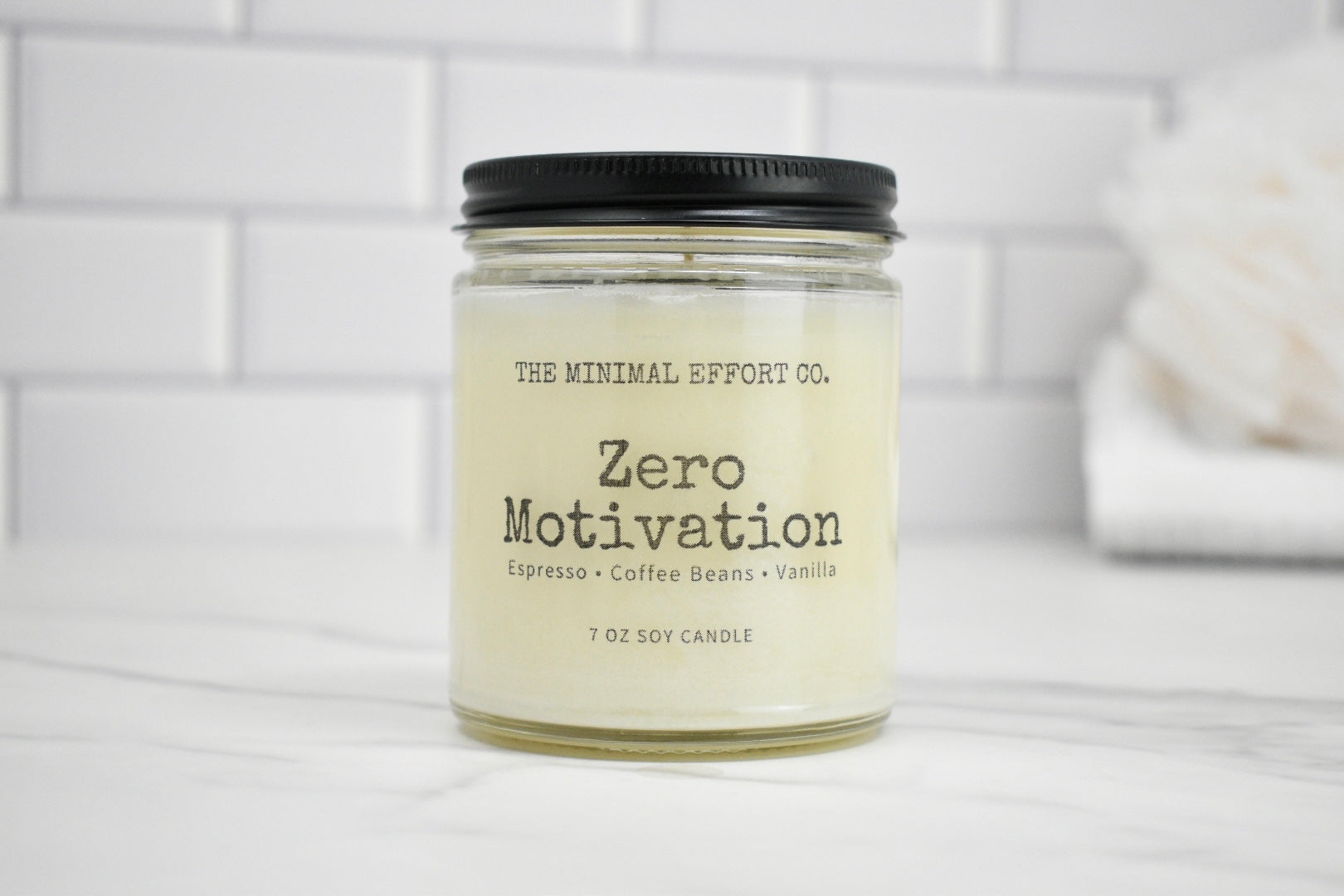 Candle jar labeled 'Zero Motivation' by The Minimal Effort Co. on a white surface with a tiled wall background.