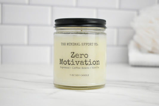 Candle jar labeled 'Zero Motivation' by The Minimal Effort Co. on a white surface with a tiled wall background.