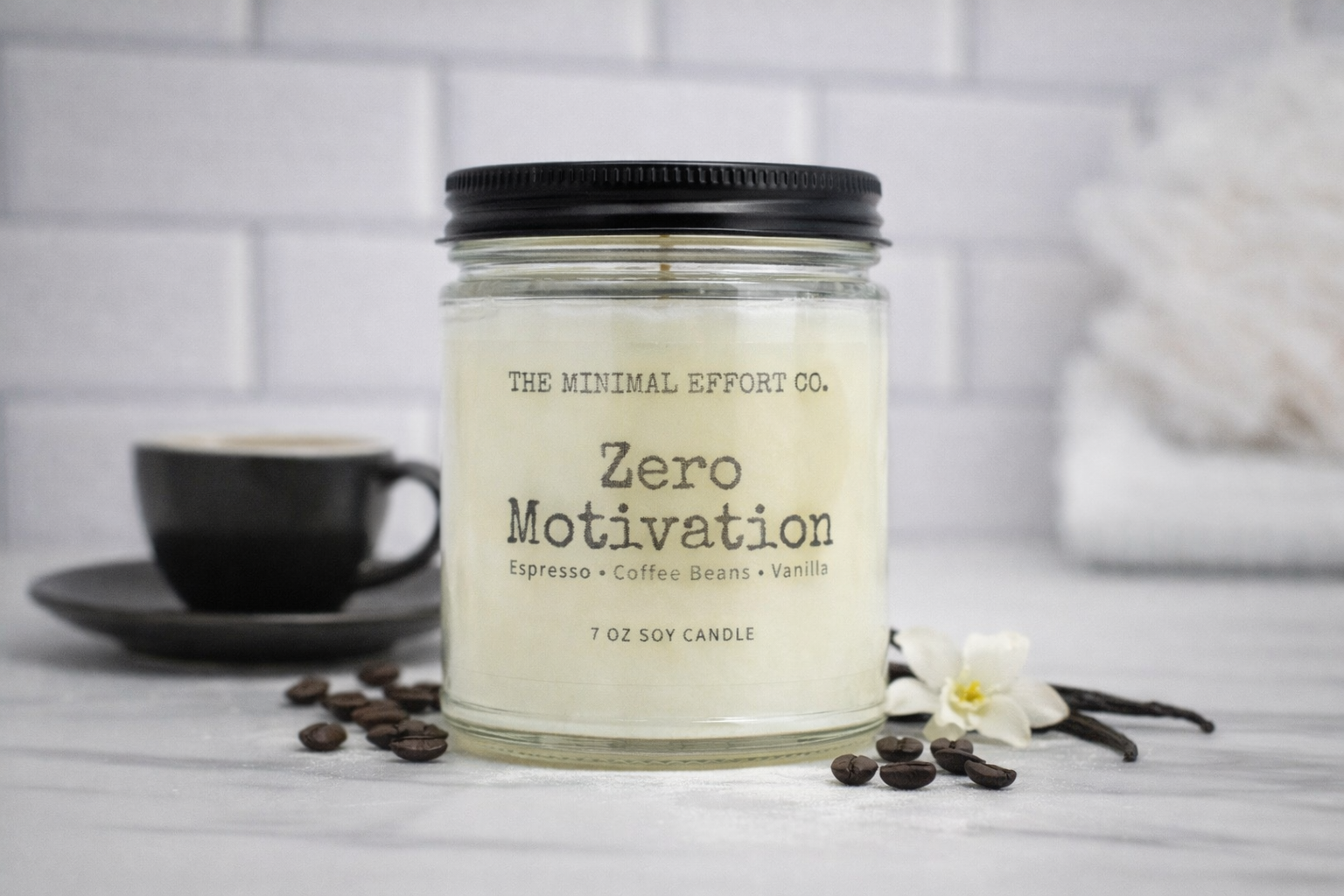 Candle labeled 'Zero Motivation' from The Minimal Effort Co. on a table with coffee beans and a cup.