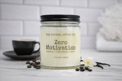 Candle labeled 'Zero Motivation' from The Minimal Effort Co. on a table with coffee beans and a cup.
