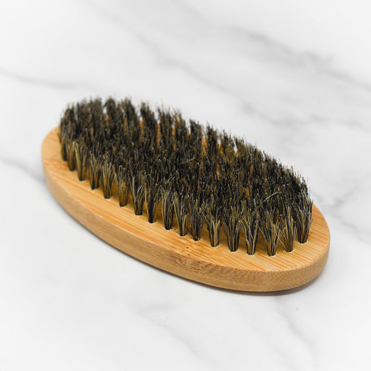 The 570 Soap Co. Beard Grooming Tool, a wooden oval beard brush featuring dense black bristles, is elegantly placed on a white marble surface, making it an essential addition to your grooming collection.